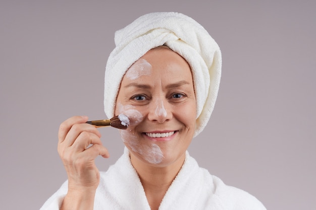 Mooie rijpe vrouw in een witte jas die een voedend masker op haar gezicht toepast. Isoleer op een grijze muur.