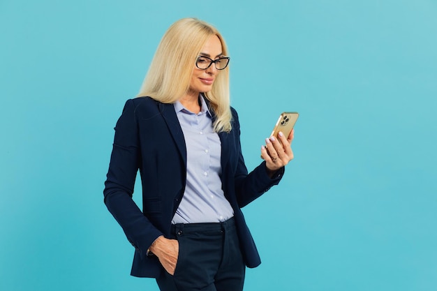 Mooie rijpe vrouw in pak en bril houdt de telefoon vast over geïsoleerde blauwe achtergrond