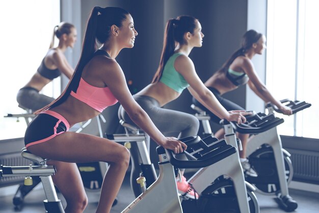 Mooie rit. Zijaanzicht van jonge mooie vrouwen met perfecte lichamen in sportkleding die wegkijken met een glimlach tijdens het fietsen in de sportschool