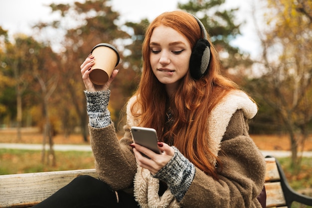 Mooie roodharige jong meisje, luisteren naar muziek met een koptelefoon zittend op een bankje, met behulp van mobiele telefoon