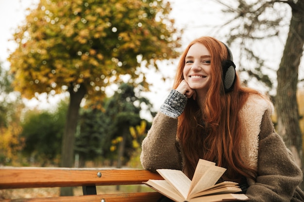 Mooie roodharige jong meisje luisteren naar muziek met koptelefoon zittend op een bankje, het lezen van een boek
