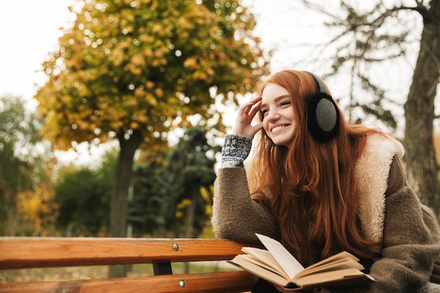 Mooie roodharige jong meisje luisteren naar muziek met koptelefoon zittend op een bankje, het lezen van een boek