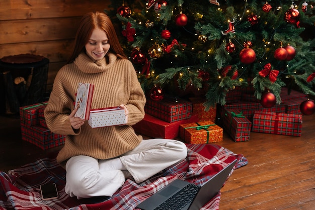 Mooie roodharige jonge vrouw die de doos van de gift van Kerstmis opent met cadeau op de achtergrond van de kerstboom