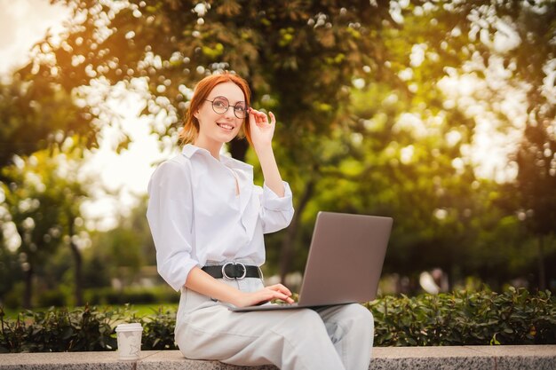 Mooie roodharige jonge vrouw zitten in het park en het gebruik van laptop student universiteit freelance slijtage...