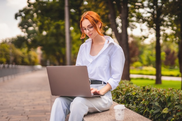 Mooie roodharige jonge vrouw zitten in het park en het gebruik van laptop student universiteit freelance slijtage...