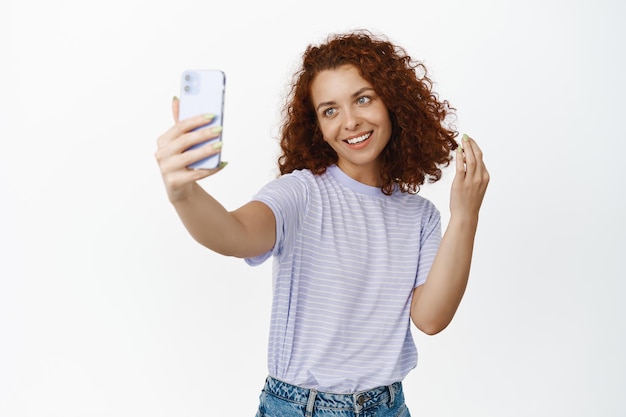 Mooie roodharige vrouw die foto's maakt, selfie maakt op mobiele telefoon, speelt met haar krullend kapsel en glimlacht, staande tegen een witte achtergrond