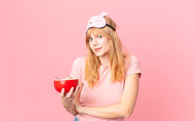 Mooie roodharige vrouw die haar schouders ophaalt, zich verward en onzeker voelt terwijl ze een pyjama draagt en een vlokkenkom vasthoudt