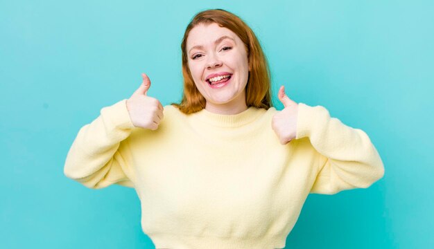 Mooie roodharige vrouw die in het algemeen lacht en er gelukkig positief zelfverzekerd en succesvol uitziet met beide duimen omhoog