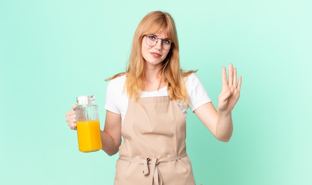 Foto mooie roodharige vrouw die lacht en er vriendelijk uitziet, met nummer vier met schort die een sinaasappelsap bereidt