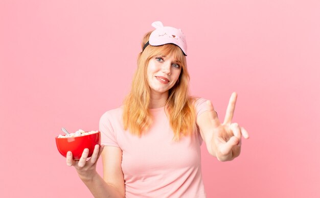 mooie roodharige vrouw die lacht en er vriendelijk uitziet, nummer twee laat zien die een pyjama draagt en een vlokkenkom vasthoudt