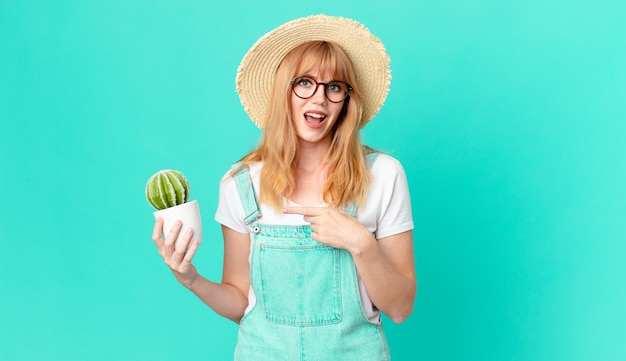 Mooie roodharige vrouw die opgewonden en verrast kijkt en naar de zijkant wijst en een ingemaakte cactus vasthoudt. boer concept