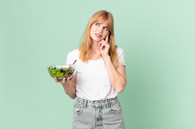 Mooie roodharige vrouw die vrolijk lacht en dagdroomt of twijfelt en een salade vasthoudt. dieet concept