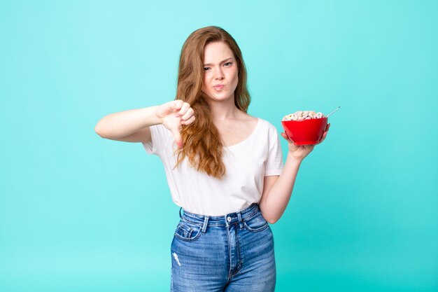 Mooie roodharige vrouw die zich boos voelt, duimen naar beneden laat zien en een ontbijtkom vasthoudt