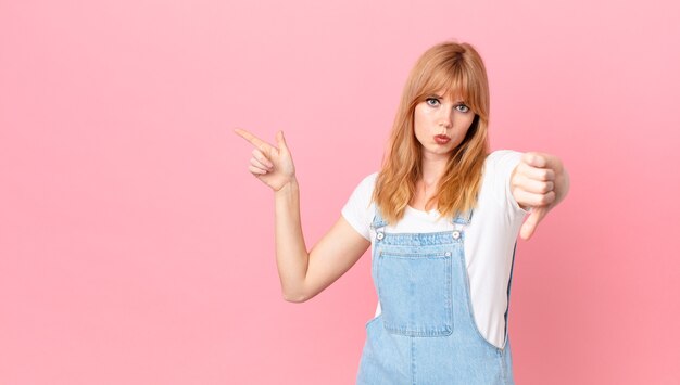 Mooie roodharige vrouw die zich boos voelt en duimen naar beneden laat zien