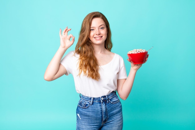 Mooie roodharige vrouw die zich gelukkig voelt, goedkeuring toont met een goed gebaar en een ontbijtkom vasthoudt