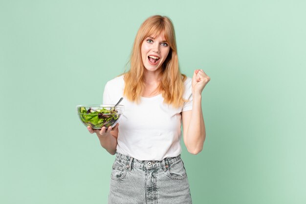 Mooie roodharige vrouw die zich geschokt voelt, lacht en succes viert en een salade vasthoudt. dieet concept