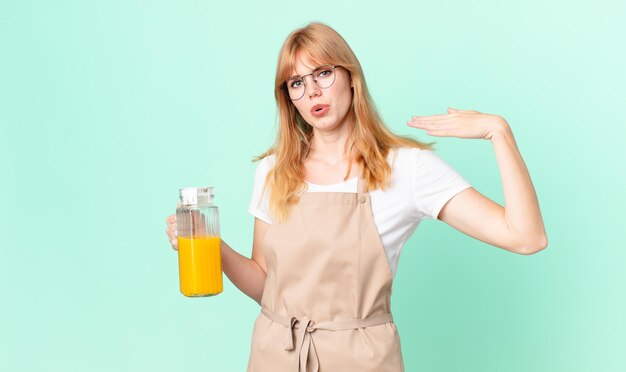 Mooie roodharige vrouw die zich gestrest, angstig, moe en gefrustreerd voelt met een schort dat een sinaasappelsap bereidt