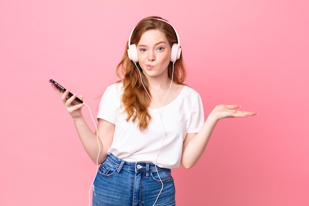 Mooie roodharige vrouw die zich verward en verward voelt en twijfelt met koptelefoon en smartphone