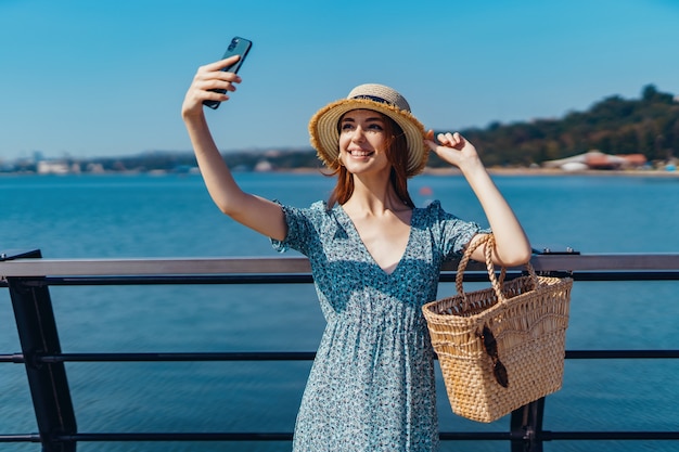 Mooie roodharige vrouw die zich voordeed op zonnige dag en selfie maakt met telefoonwandelingen langs de pier in de buurt van de...