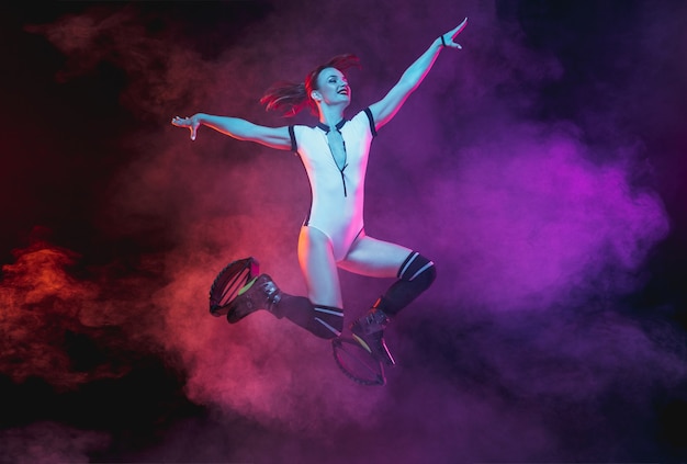 Mooie roodharige vrouw in sportkleding springen in een kangoo springt schoenen geïsoleerd op donkere achtergrond met kleurovergang studio in neon verlichte rook. actieve beweging, actie, fitness en wellness. slank model.