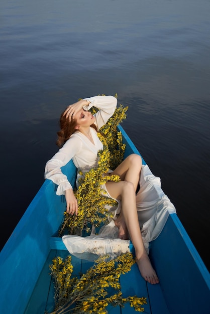 Mooie roodharige vrouw ligt in boot met bloemen in haar handen. Schoonheid romantisch portret meisje in lange witte jurk in houten boot op lake