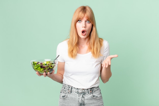 Foto mooie roodharige vrouw verbaasd, geschokt en verbaasd met een ongelooflijke verrassing en met een salade. dieet concept