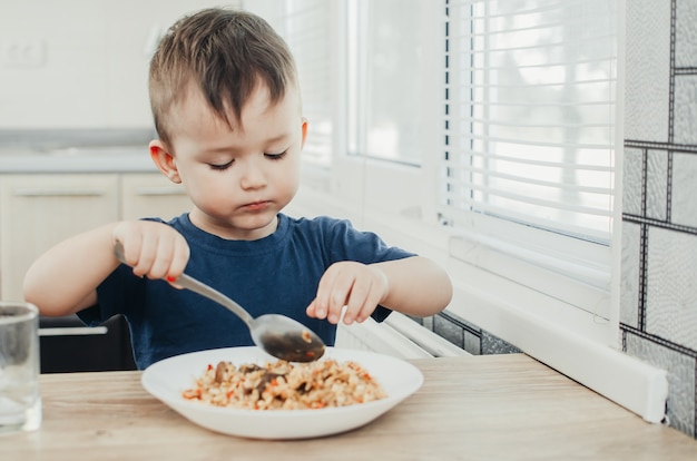 Mooie schattige baby eet rijst met een lepel in de keuken