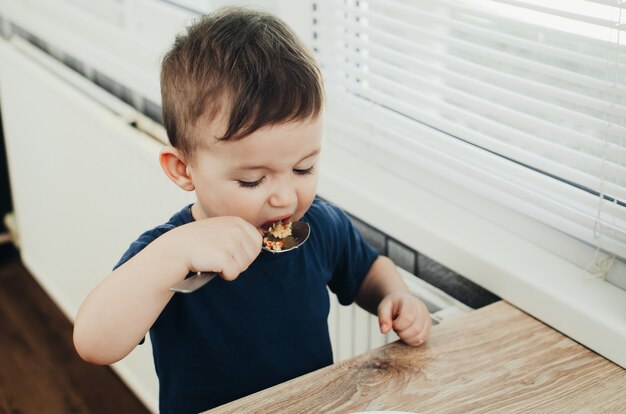 Mooie schattige baby eet rijst met een lepel in de keuken