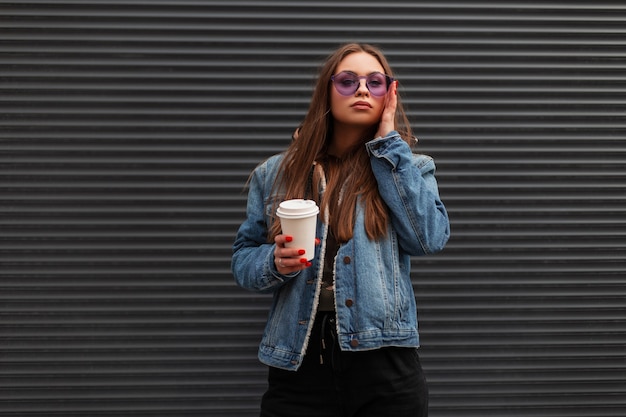 Mooie schattige jonge hipster vrouw in modieuze denim blauwe jas in trendy paarse bril met kop warme koffie poseert in de buurt van een grijze muur in de stad. Stedelijk sexy meisje poseren model op straat.