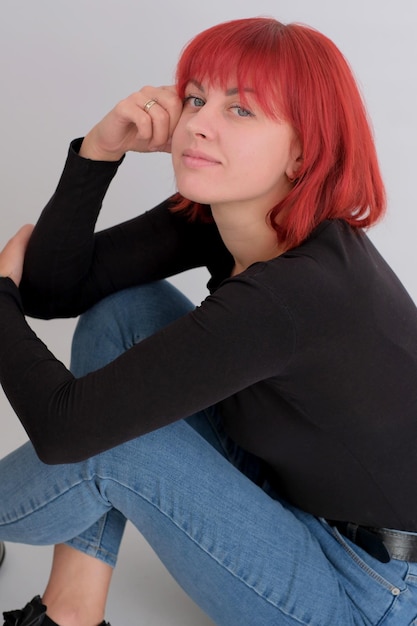 Mooie schattige jonge vrouw met oranje kapsel in spijkerbroek zitten poseren in studio op witte achtergrond