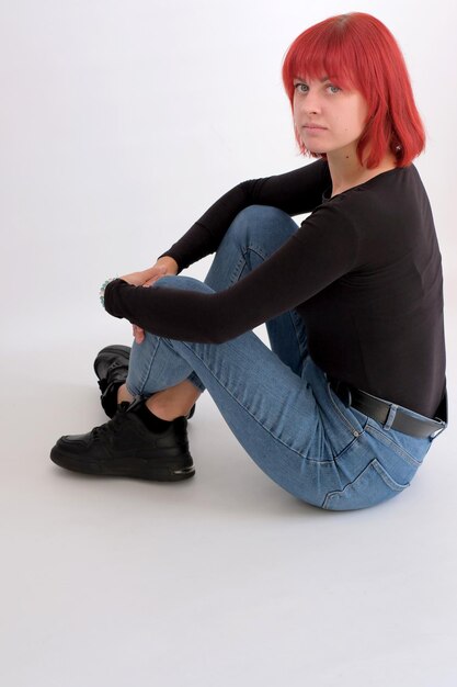 Mooie schattige jonge vrouw met oranje kapsel in spijkerbroek zitten poseren in studio op witte achtergrond