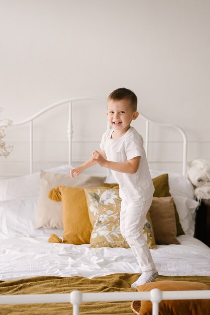 Mooie schattige vierjarige jongen in witte kleren glimlacht en springt op het bed op de lichte achtergrond van het huis