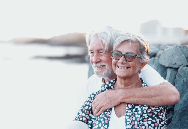 Mooie senior paar knuffelen glimlachend in de buitenlucht op zee wegkijken. Kaukasisch paar met wit haar dat geniet van pensioen en zonsonderganglicht