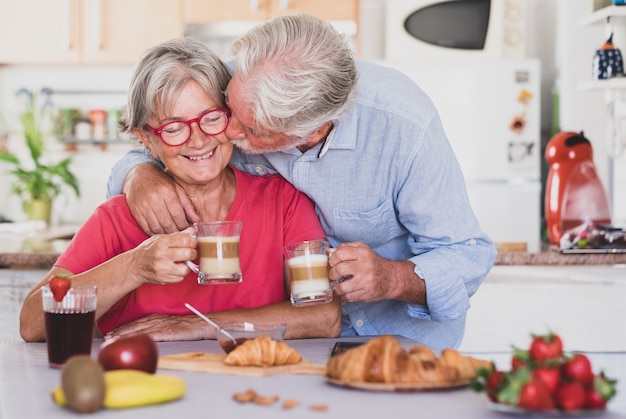 Mooie senior paar ontbijten thuis, 's ochtends beginnen met een kus. Warme cappuccino in de handen