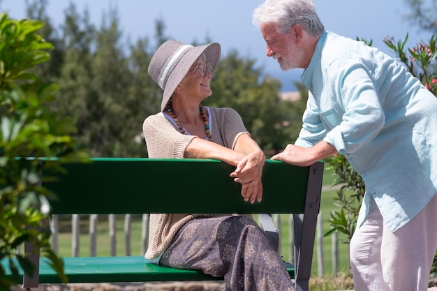 Mooie senior paar zitten buiten op een bankje van openbaar park kijken in de ogen Lachende bejaarde grootouders genieten van ontspannen en pensioen op zonnige dag