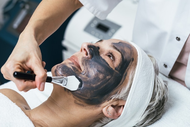 Mooie senior vrouw die een gezichtsmasker van klei ontvangt met verjongende effecten in de schoonheidssalon van de spa.