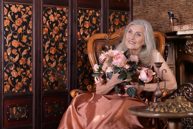 Mooie senior vrouw in gouden jurk in vintage stoel met bloemen