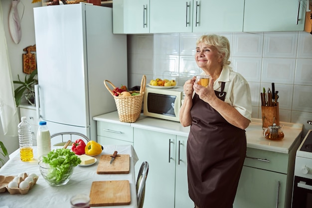 Mooie senior vrouw in schort die thuis thee drinkt