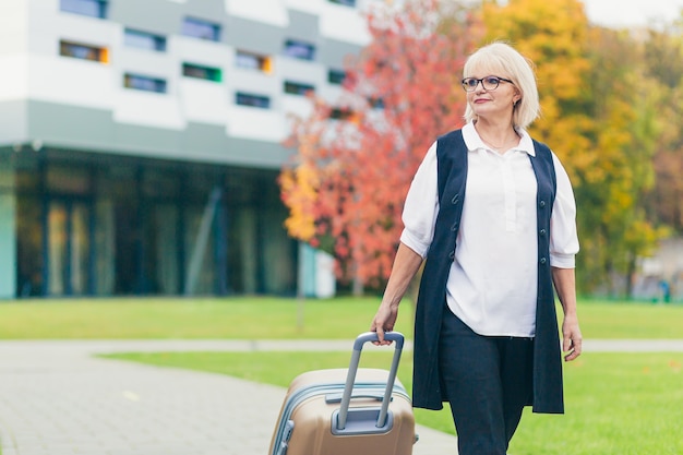 Mooie senior vrouw reist met een grote reiskoffer, op de achtergrond van een prachtig modern gebouw