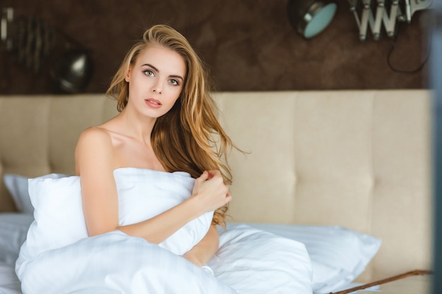 Mooie sensuele jonge vrouw zittend op bed gewikkeld in wit dekbed