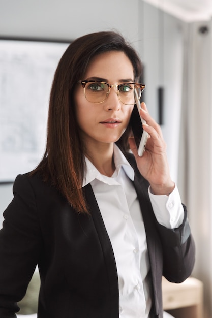 mooie serieuze jonge zakenvrouw in formele kleding binnenshuis thuis praten via de mobiele telefoon.