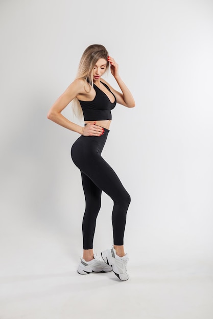 Mooie sexy fitness model blonde vrouw met een slank fit lichaam in een modieuze zwarte sportkleding met legging sneakers en een top op een witte achtergrond