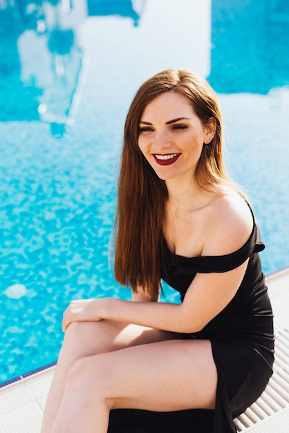 Mooie sexy jonge vrouw in zwarte jurk rusten bij het blauwe zwembad en lachen