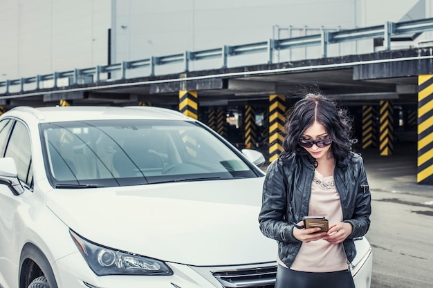 Mooie sexy vrouwelijke model in lederen kleding met een witte auto en een smartphone op de parkeerplaats