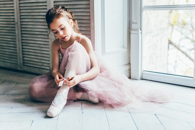 Mooie sierlijke ballerina in roze tutu rok zet pointe-schoenen in de buurt van groot raam in witte lichte zaal