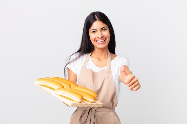 Mooie Spaanse chef-kokvrouw die trots is, positief glimlacht met duimen omhoog en een dienblad met broodbroodjes vasthoudt