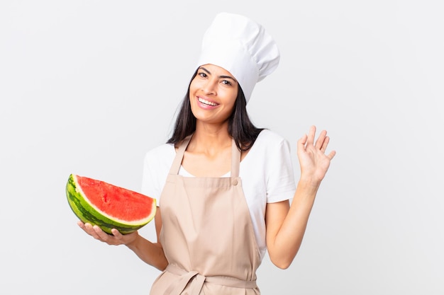 Mooie Spaanse chef-kokvrouw die vrolijk lacht, met de hand zwaait, je verwelkomt en begroet en een watermeloen vasthoudt