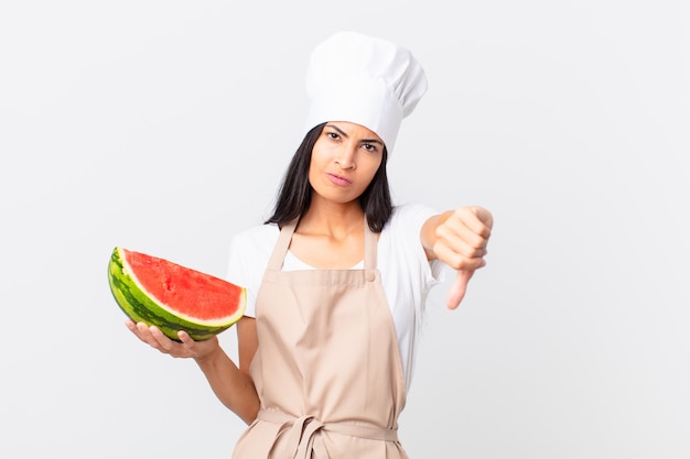 Mooie Spaanse chef-kokvrouw die zich boos voelt, duimen naar beneden laat zien en een watermeloen vasthoudt