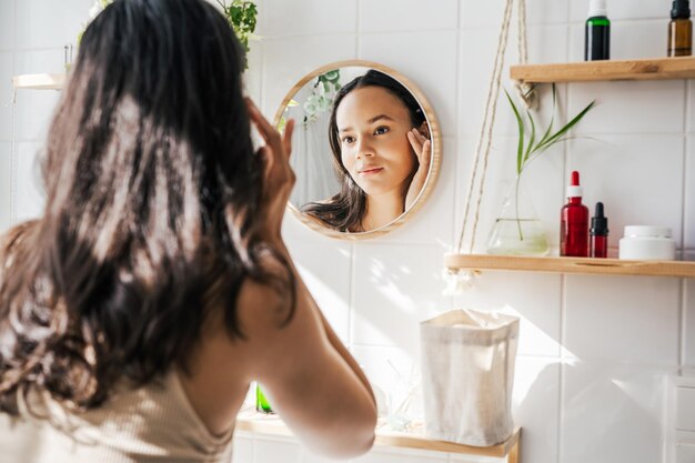 Mooie Spaanse vrouw die in de spiegel kijkt in een witte milieuvriendelijke badkamer Houten planken en herbruikbare cosmetische flessen Wellness huidverzorgingsconcept