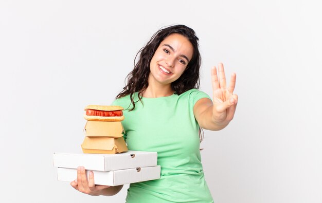 Mooie Spaanse vrouw die lacht en er vriendelijk uitziet, nummer drie laat zien en fastfooddozen vasthoudt